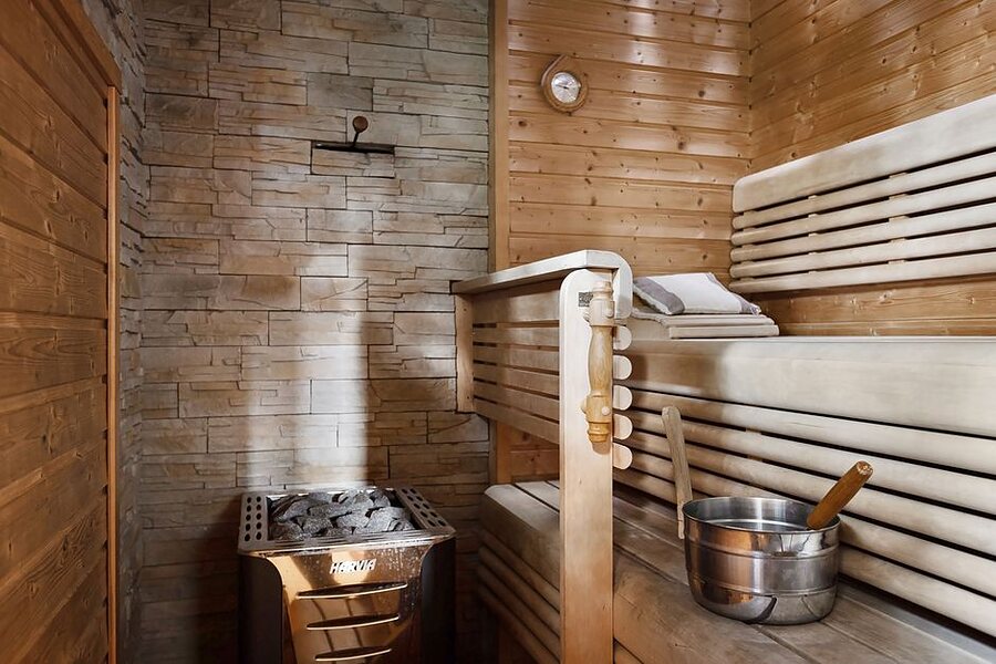 A wooden sauna interior with benches, a stone heater and a water bucket