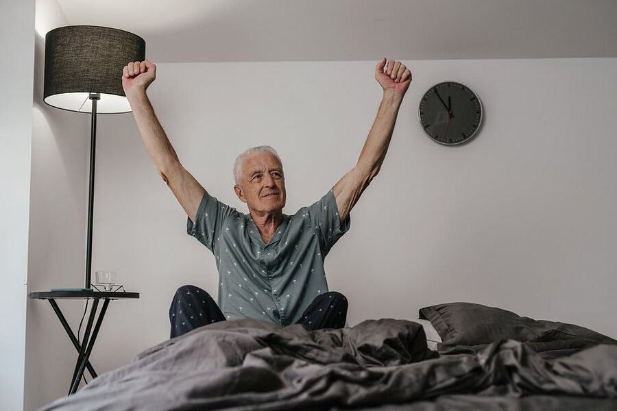 An older man sitting on the edge of his bed, arms stretched overhead